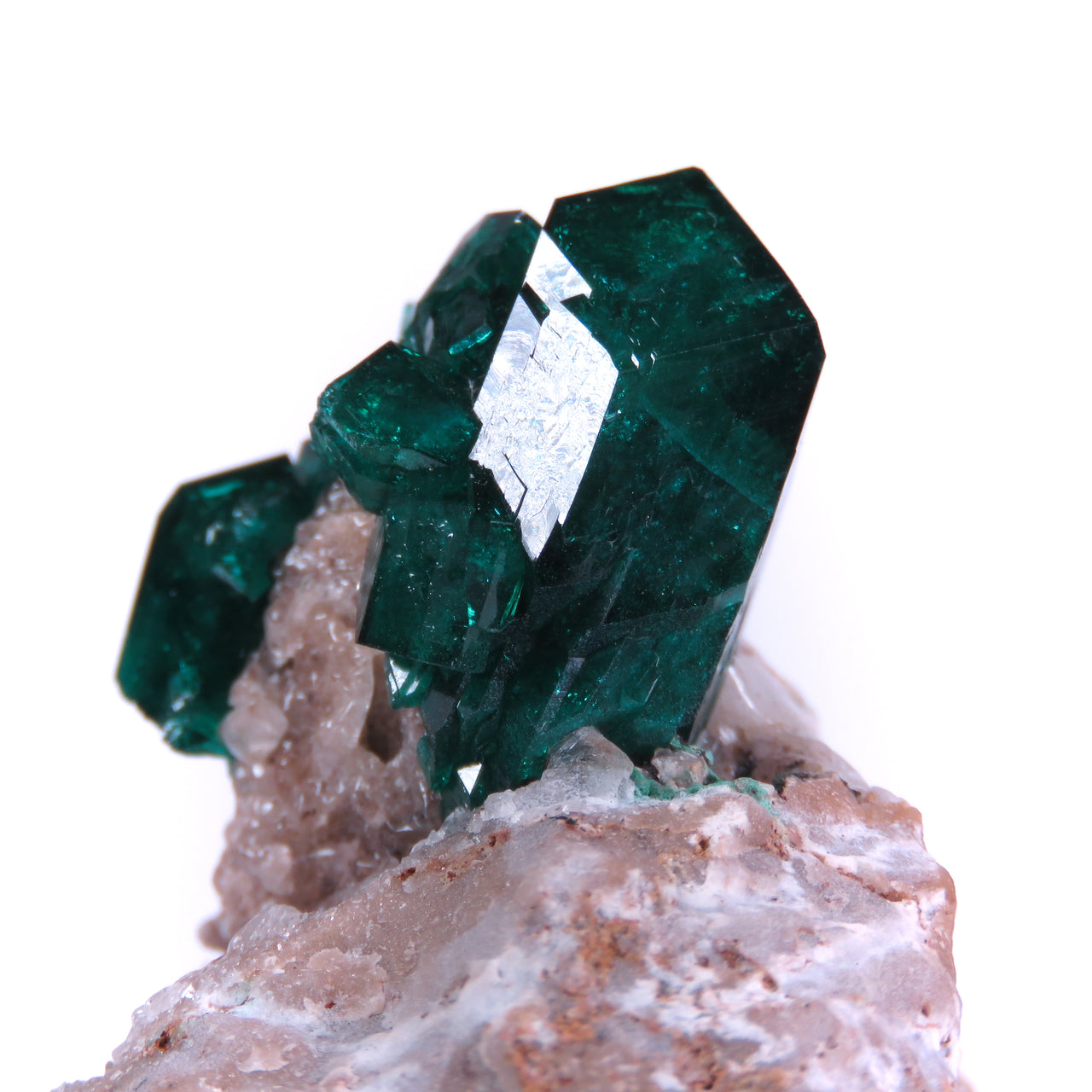 Two views of a mineral specimen featuring deep emerald-green dioptase crystals perched on pale pink-brown quartz matrix. The dioptase forms a cluster of well-formed, prismatic crystals with the largest crystal reaching nearly an inch in length. The crystals are highly translucent with glassy luster, showing bright reflective faces, particularly visible in the second, closer view. The main crystal displays perfect geometric form, rising dramatically from the rough, textured quartz base.