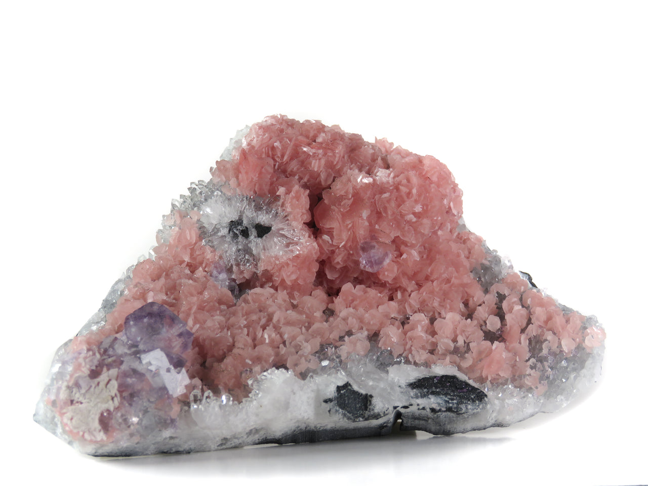 A delightful rhodochrosite specimen featuring flattened rhombohedral crystals with soft pink coloration, accompanied by purple fluorite and crystalline quartz matrix. The 12 cm specimen from China's Wutong Mine displays classic three-species paragenesis, pyrite-dusted crystal edges, and exhibits striking UV fluorescence in the fluorite crystals.